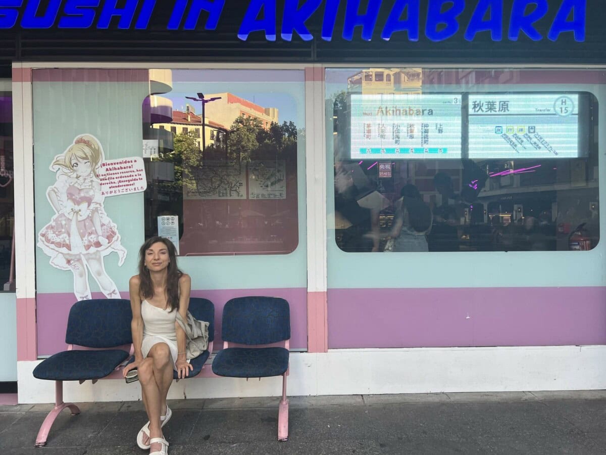 A woman sits on a bench outside a sushi restaurant in Akihabara, with anime-themed decor and a transit map visible in the window.