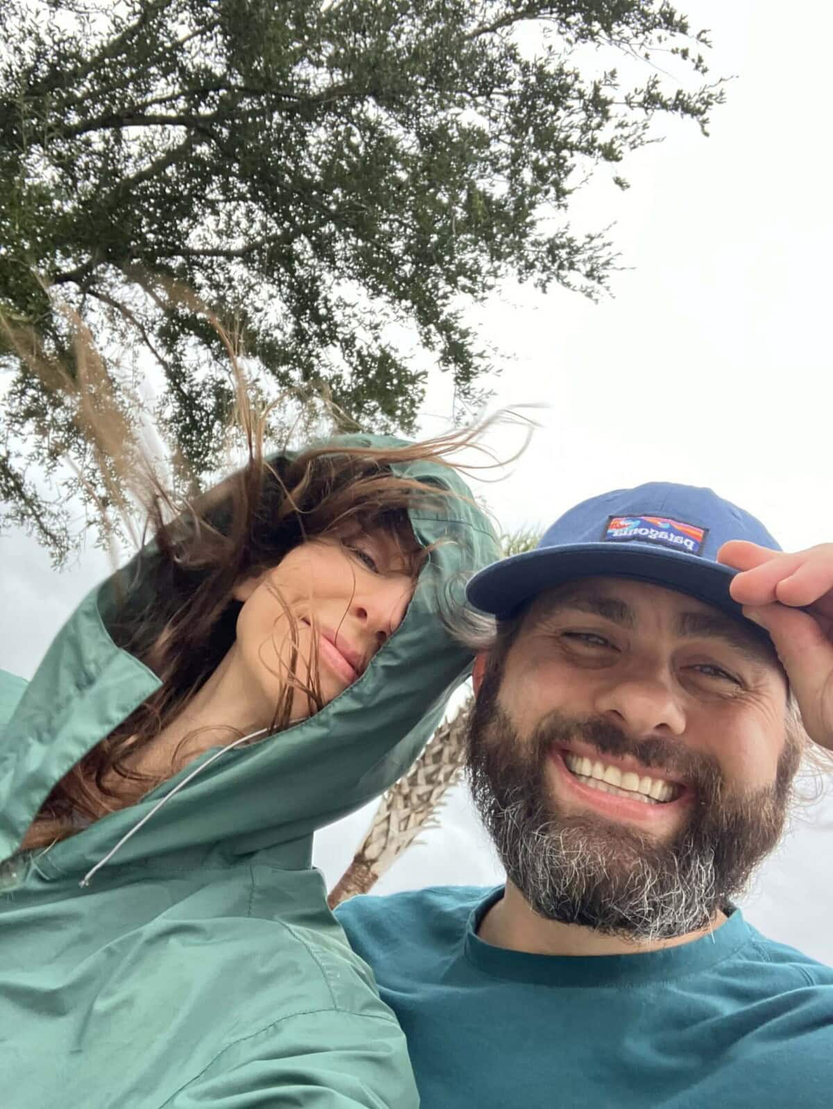 A woman in a green jacket stands next to a smiling man in a blue cap. It appears windy, as her hair is blown across her face. Trees and a cloudy sky are in the background.