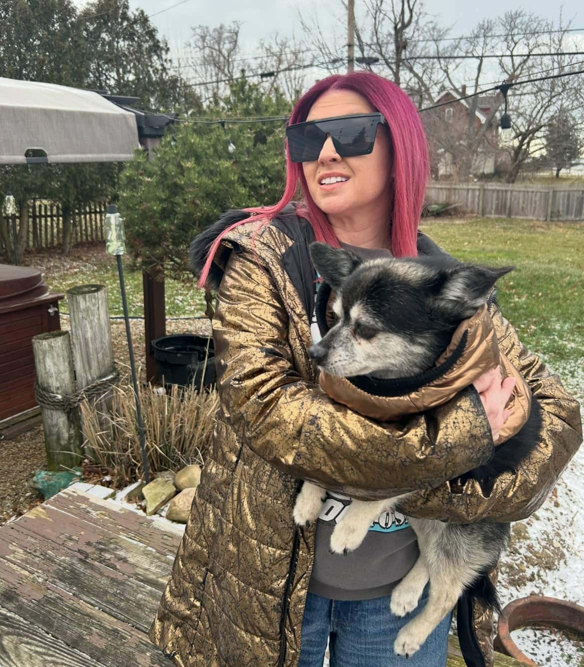 A woman with pink hair and sunglasses holds a black and white dog outside in a backyard on a cloudy day.