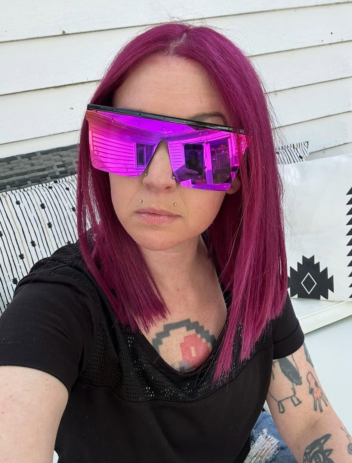 A person with straight magenta hair and large reflective pink sunglasses sits outside against a white wall, wearing a black shirt and displaying visible tattoos.