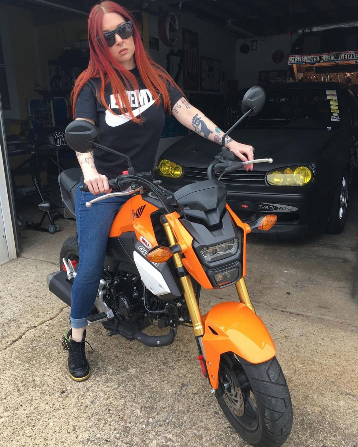 A woman with red hair wearing sunglasses sits on an orange Honda motorcycle in front of a garage, with cars visible in the background.