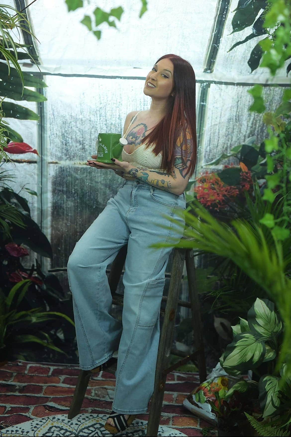 A woman with long red hair sits on a wooden stool in a greenhouse, holding a green mug. She wears a white top, light blue jeans, and sandals, surrounded by lush plants and flowers.