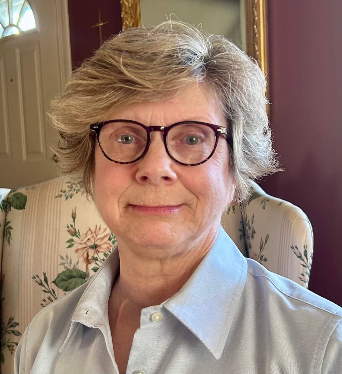 An older woman with short gray hair and glasses sits on a floral armchair, wearing a light blue collared shirt, in a warmly lit room.