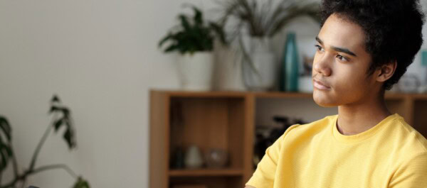A young person wearing a yellow shirt looks thoughtfully to the side, sitting indoors with plants and shelves in the background.