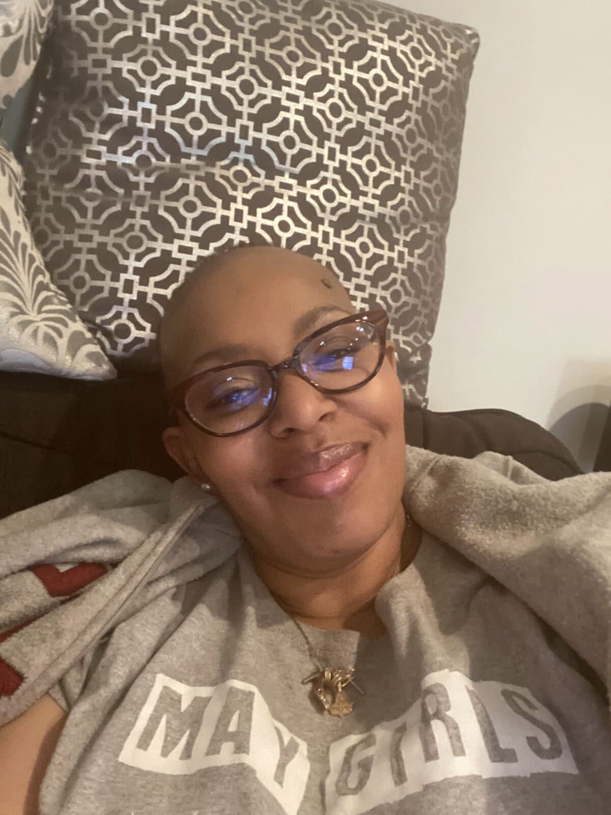 A person wearing glasses and a "May Girls" shirt smiles while sitting on a couch with patterned pillows behind them.