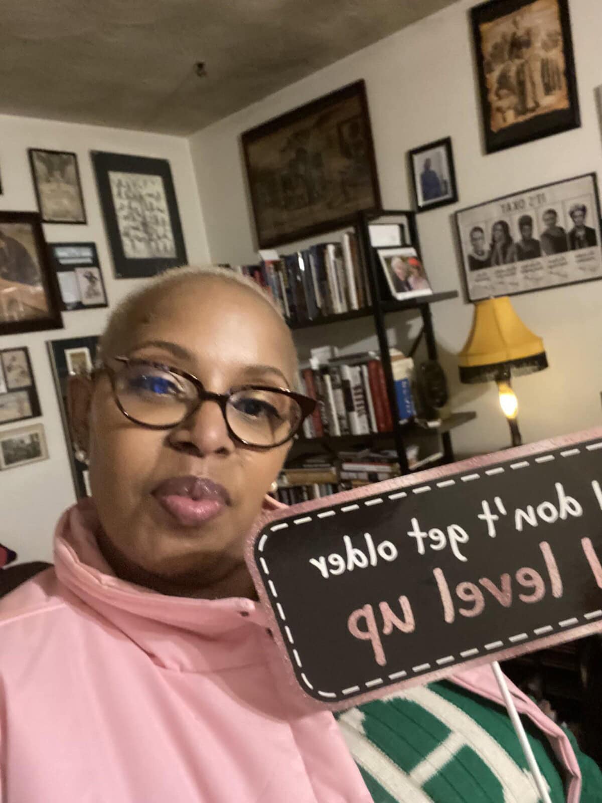 A woman wearing glasses and a pink jacket holds a sign that says "I don't get older, I level up" in a room with bookshelves, framed photos, and wall art.