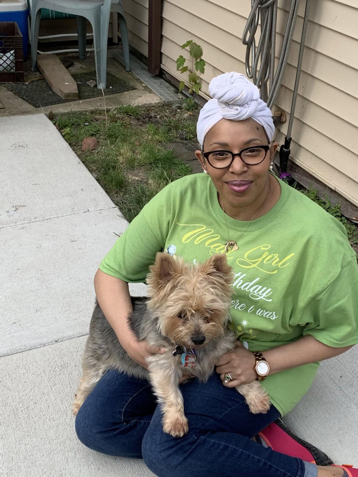 A woman wearing glasses and a green shirt sits on the ground outside, holding a small dog on her lap.