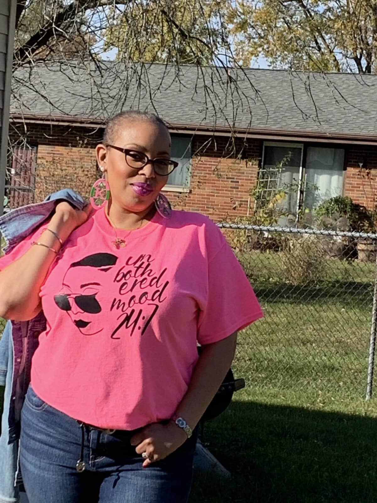 A woman wearing glasses and a bright pink T-shirt that reads "I'm both tired and mood 24:7" stands outside in front of a brick house and a chain-link fence.