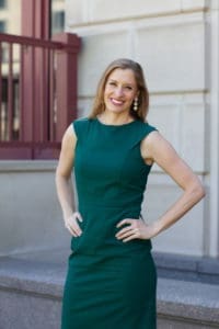 Woman in a sleeveless green dress stands outdoors with hands on hips, smiling at the camera in front of a stone building.