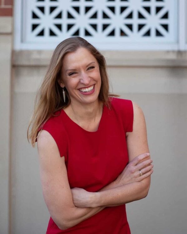 Woman with long light brown hair wearing a sleeveless red dress stands smiling with arms crossed in front of a light-colored wall and decorative window.