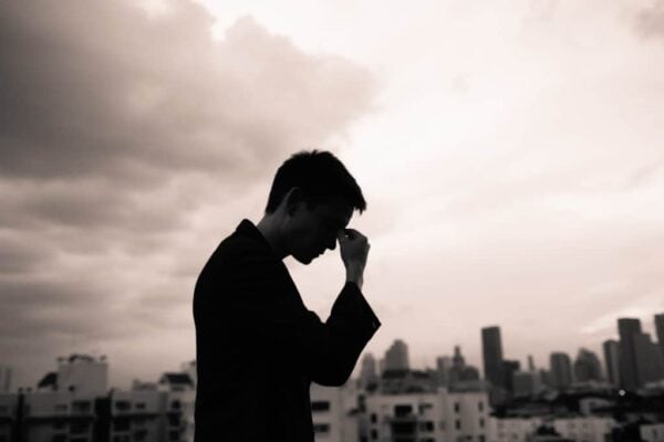 A person stands in silhouette on a rooftop with a city skyline in the background, holding their head as if deep in thought.