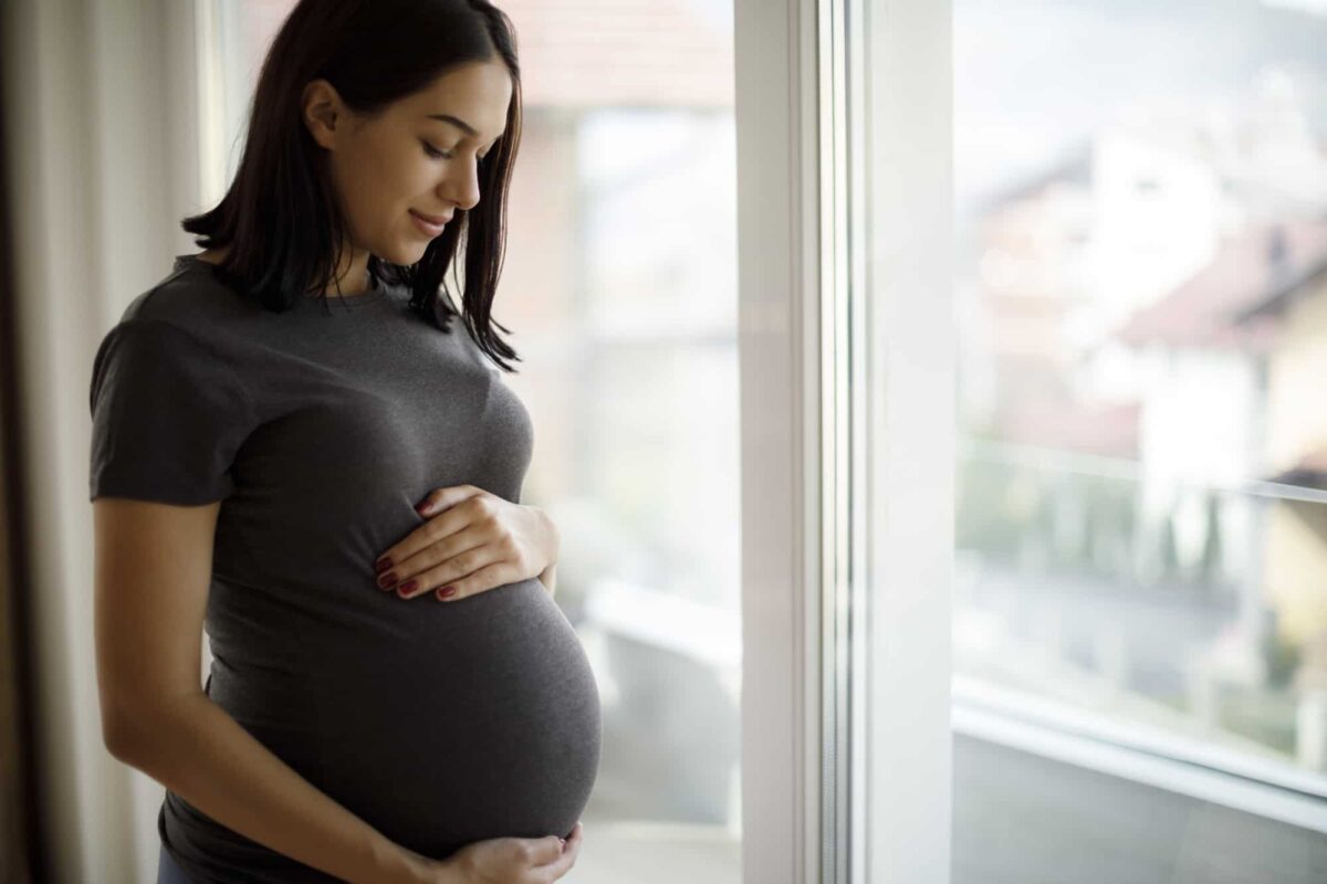 A pregnant woman in a grey t-shirt stands by a window, gently holding her belly and looking down with a slight smile.