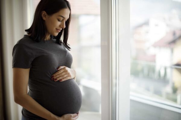 A pregnant woman in a grey t-shirt stands by a window, gently holding her belly and looking down with a slight smile.