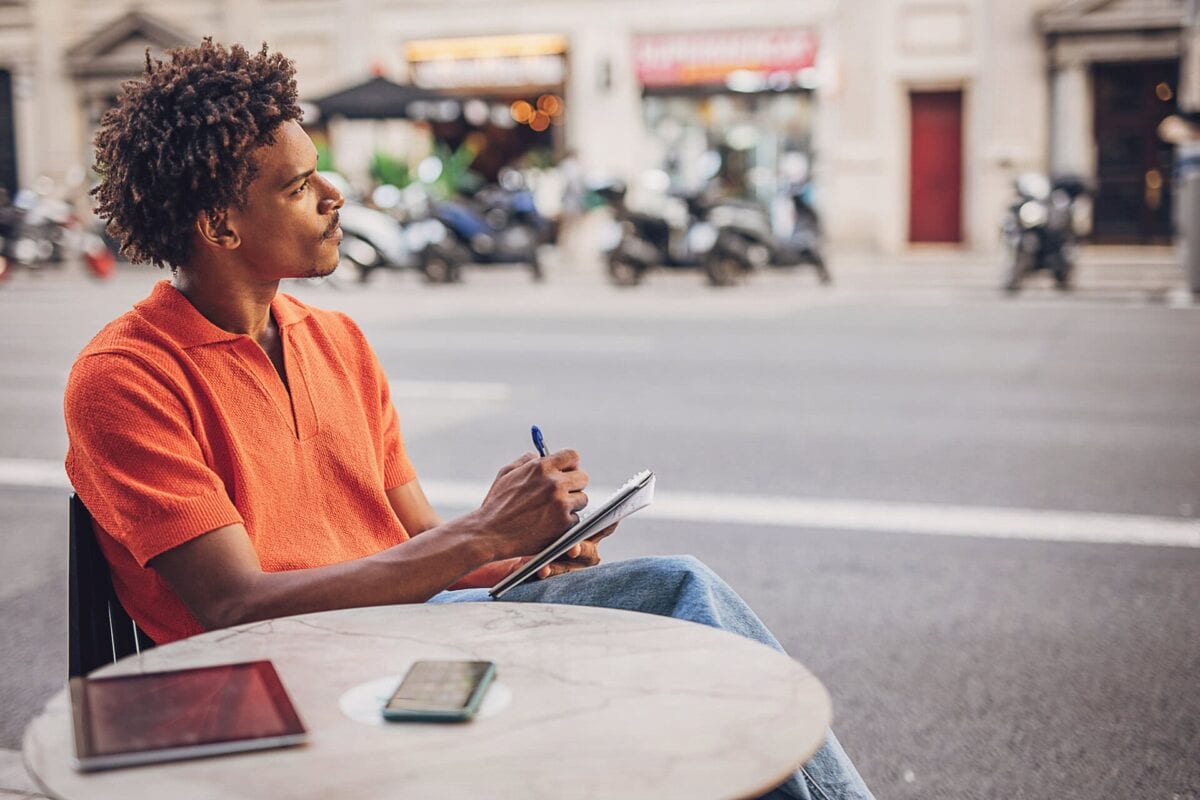 A person sits at an outdoor table with a tablet and phone, writing in a notebook while looking thoughtfully across the street.