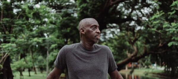 A man in a gray t-shirt stands outdoors with trees and greenery in the background, looking to the side.