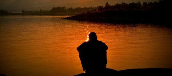 A person sits by the edge of a calm body of water at sunset, with their silhouette visible against the orange reflections on the water.