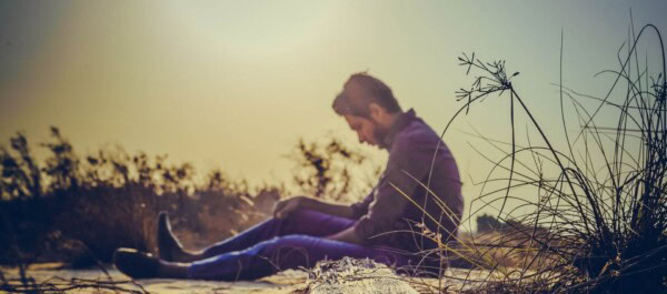 A man sits on the ground outdoors in sunlight, looking down with his arms resting on his knees and plants around him.