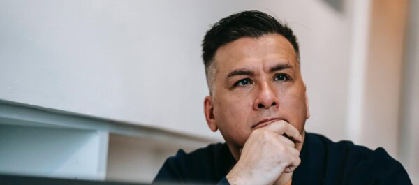 Man with short dark hair and a navy shirt resting his chin on his hand, looking thoughtfully ahead in an indoor setting.