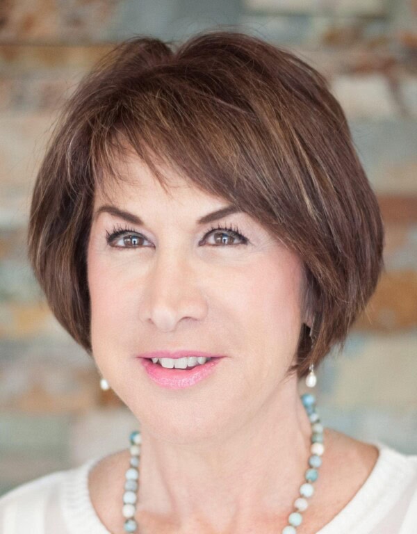 A woman with short brown hair and light makeup, wearing a white top and a beaded necklace, poses in front of a blurred background.
