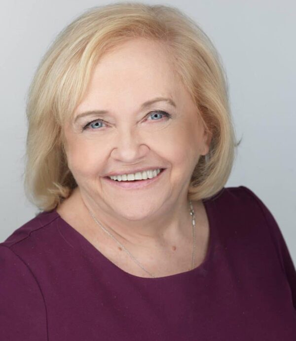 An older woman with short blond hair and blue eyes smiles at the camera, wearing a purple top and a silver necklace against a plain light background.