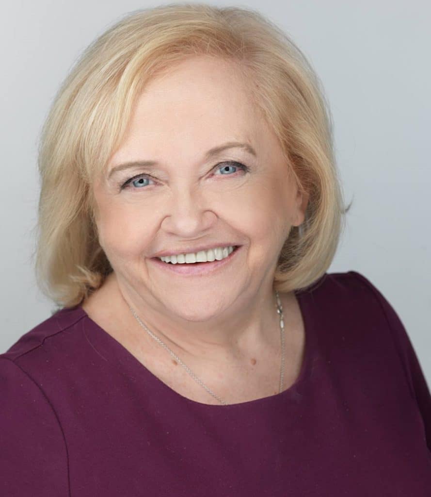 An older woman with short blond hair and blue eyes smiles at the camera, wearing a purple top and a silver necklace against a plain light background.