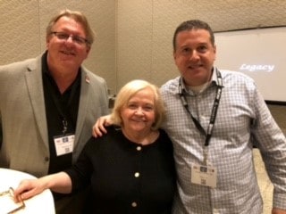Three adults, two men and one woman, pose together indoors at an event wearing conference badges. A projection screen in the background displays the word "Legacy.
