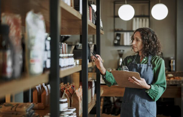 A woman in an apron holds a clipboard and pen while checking inventory on shelves in a store.