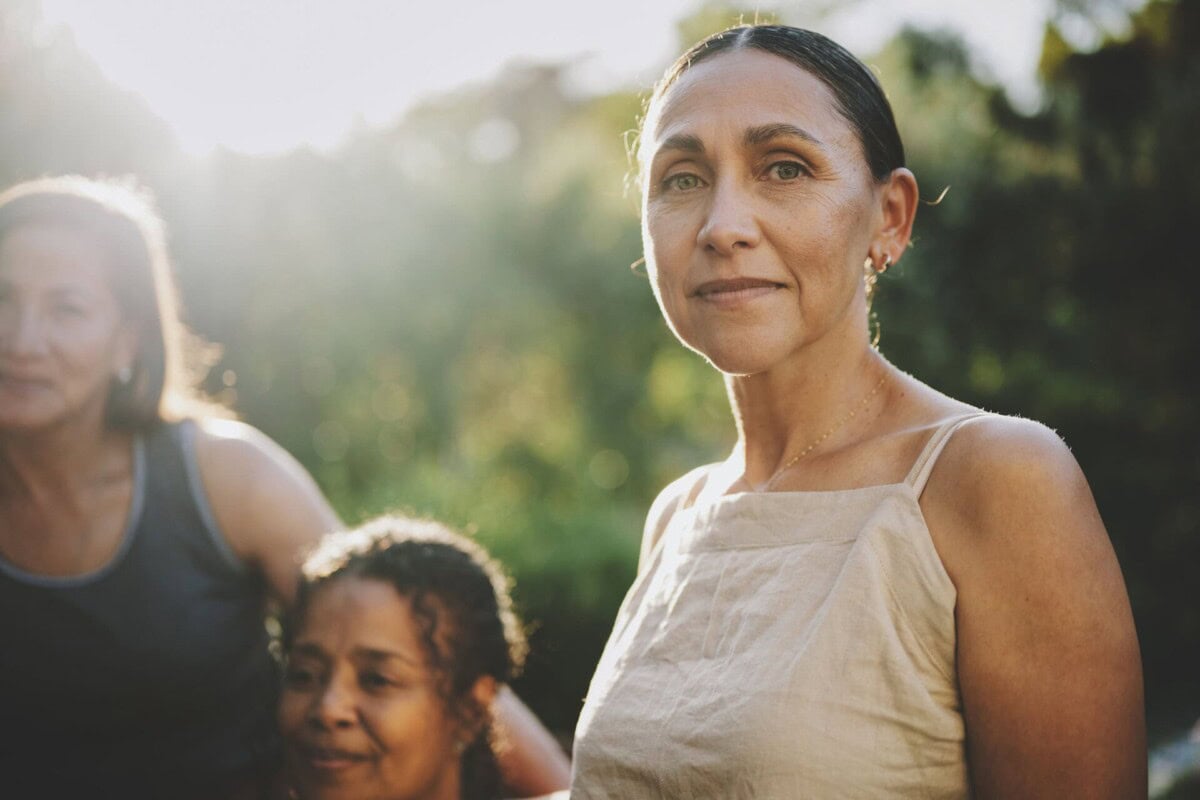 Three women stand outdoors in sunlight, with greenery in the background. One woman in front looks directly at the camera while the others are slightly behind her and to the left.