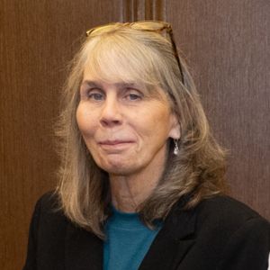 A woman with long gray hair, wearing glasses on her head, a teal top, and a black blazer, stands in front of a wooden background.