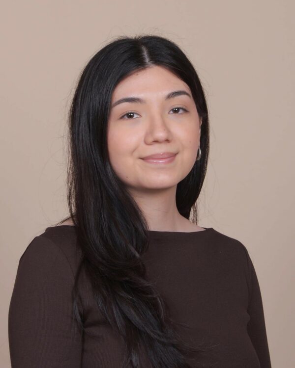 A woman with long black hair and a brown top smiles slightly while posing in front of a plain beige background.
