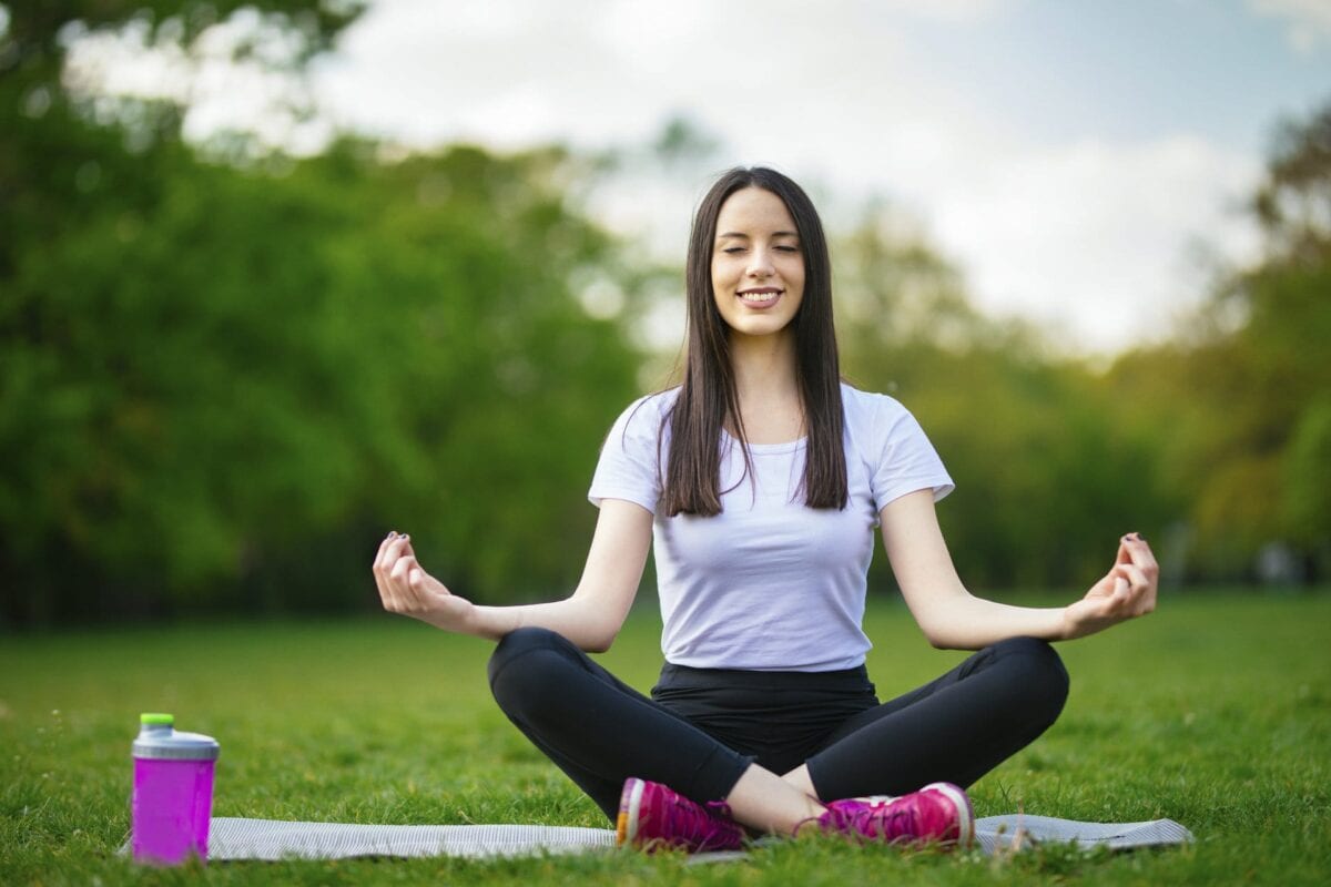A woman sits cross-legged on a yoga mat outdoors, meditating with her eyes closed and hands resting on her knees. A water bottle is placed beside her on the grass.