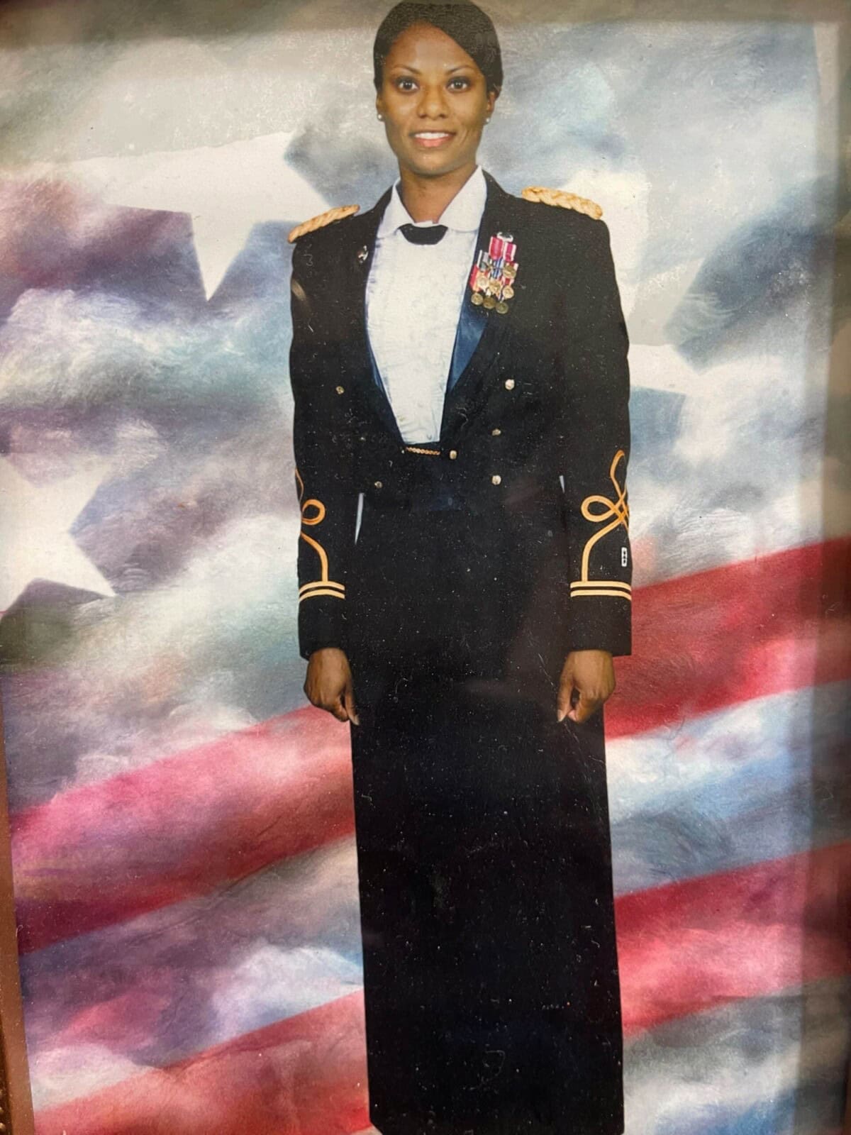 A woman in a formal military dress uniform stands in front of a backdrop featuring the American flag.