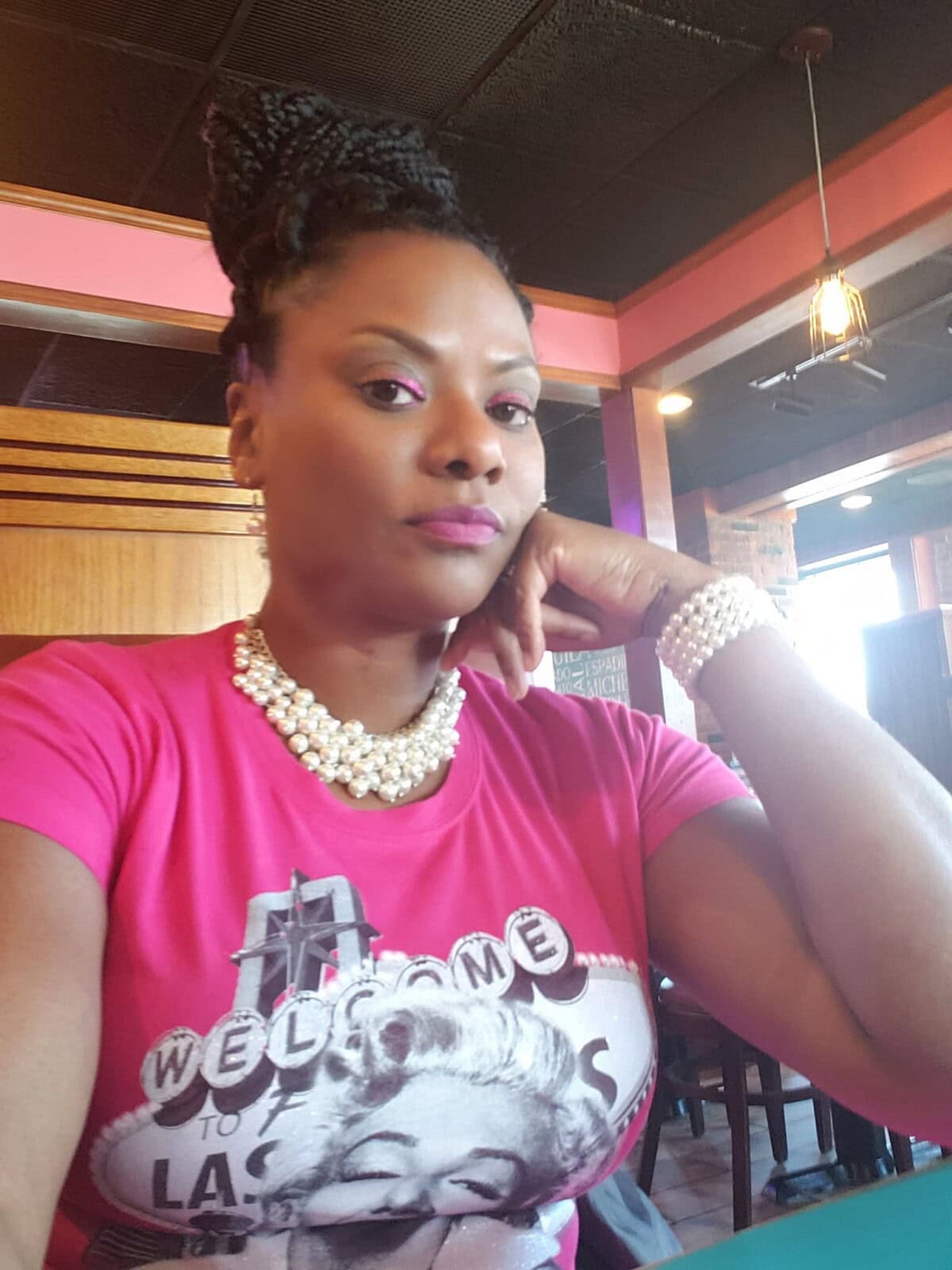 A woman wearing a pink "Welcome to Las Vegas" T-shirt and pearl jewelry sits indoors, resting her chin on her hand, looking at the camera.