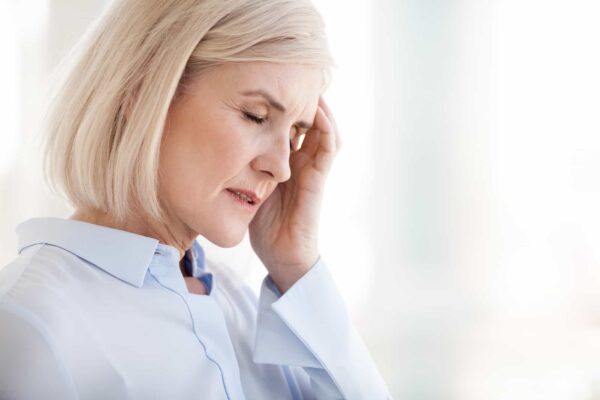Older woman with short blonde hair closes her eyes and touches her temple, appearing to experience discomfort or a headache, while wearing a light blue blouse.
