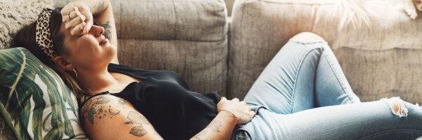 A woman with tattoos lies on a couch, wearing a headband and ripped jeans, resting with her hand on her forehead and eyes closed.