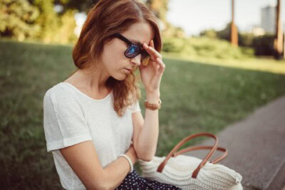 A woman wearing sunglasses sits on grass near a sidewalk, touching her forehead with one hand and holding a bag in her lap.