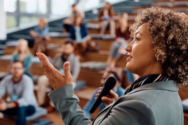 A person holding a microphone speaks to an audience seated in a tiered lecture hall.