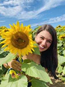 A woman with long dark hair smiles while standing in a sunflower field, holding a large sunflower, under a blue sky with scattered clouds.