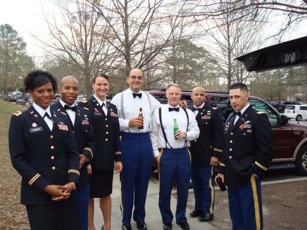 A group of people in military dress uniforms stand together outdoors, some holding drinks, with trees and parked vehicles in the background.
