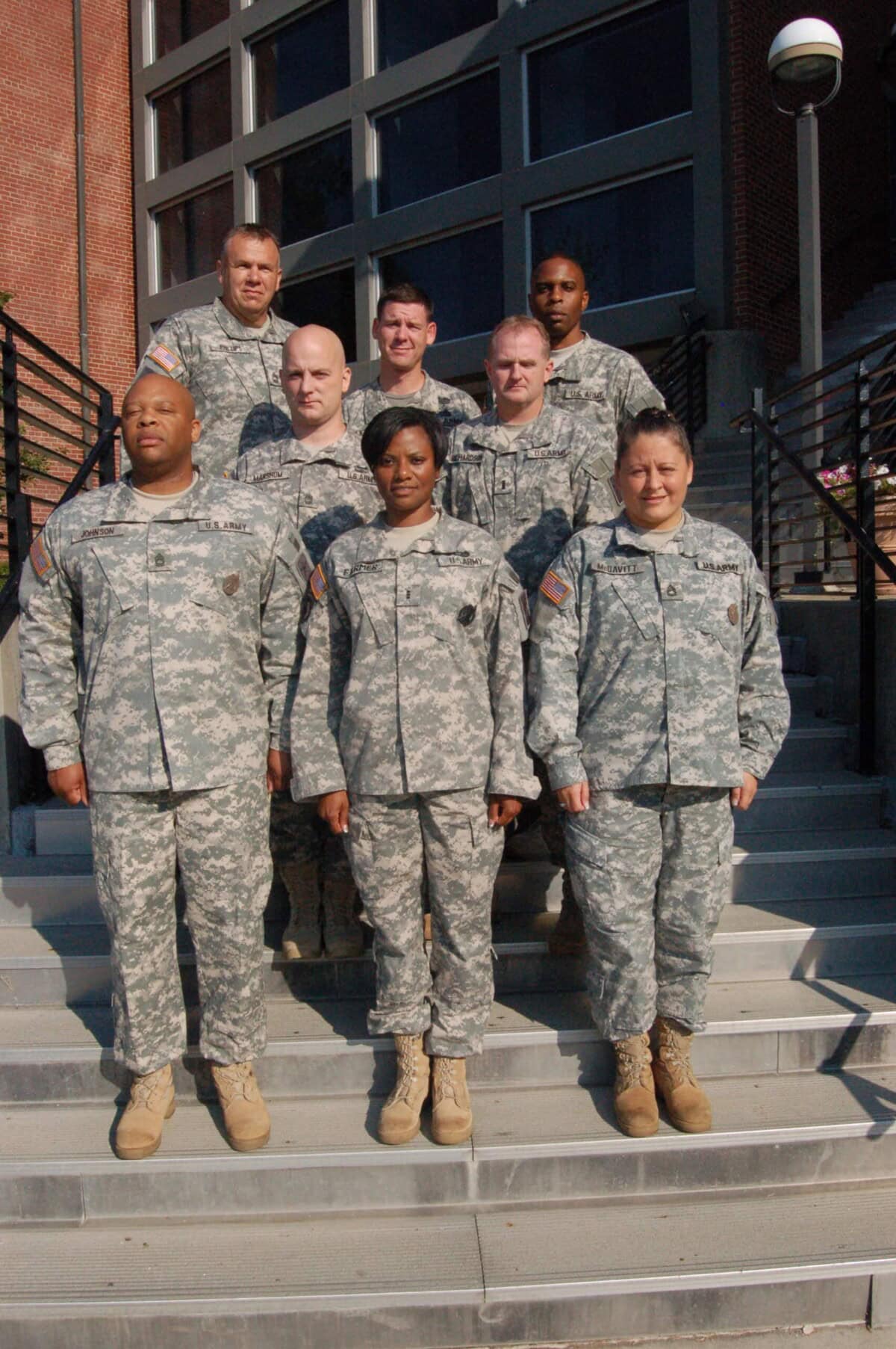 Seven soldiers in camouflage uniforms stand on outdoor steps in two rows in front of a brick building, posing for a group photo.