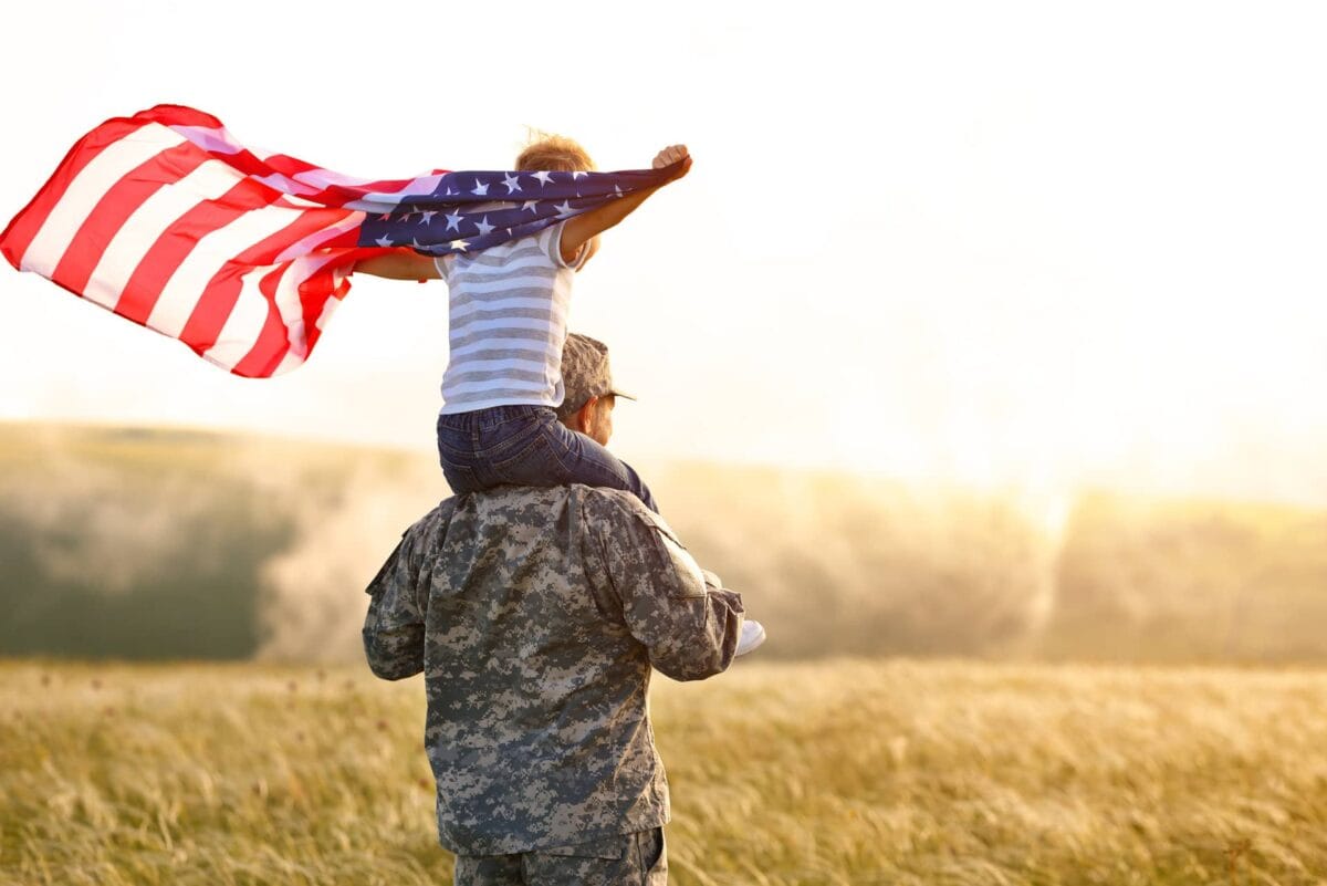 A person in military uniform stands in a field with a child on their shoulders; the child holds an American flag draped behind them.
