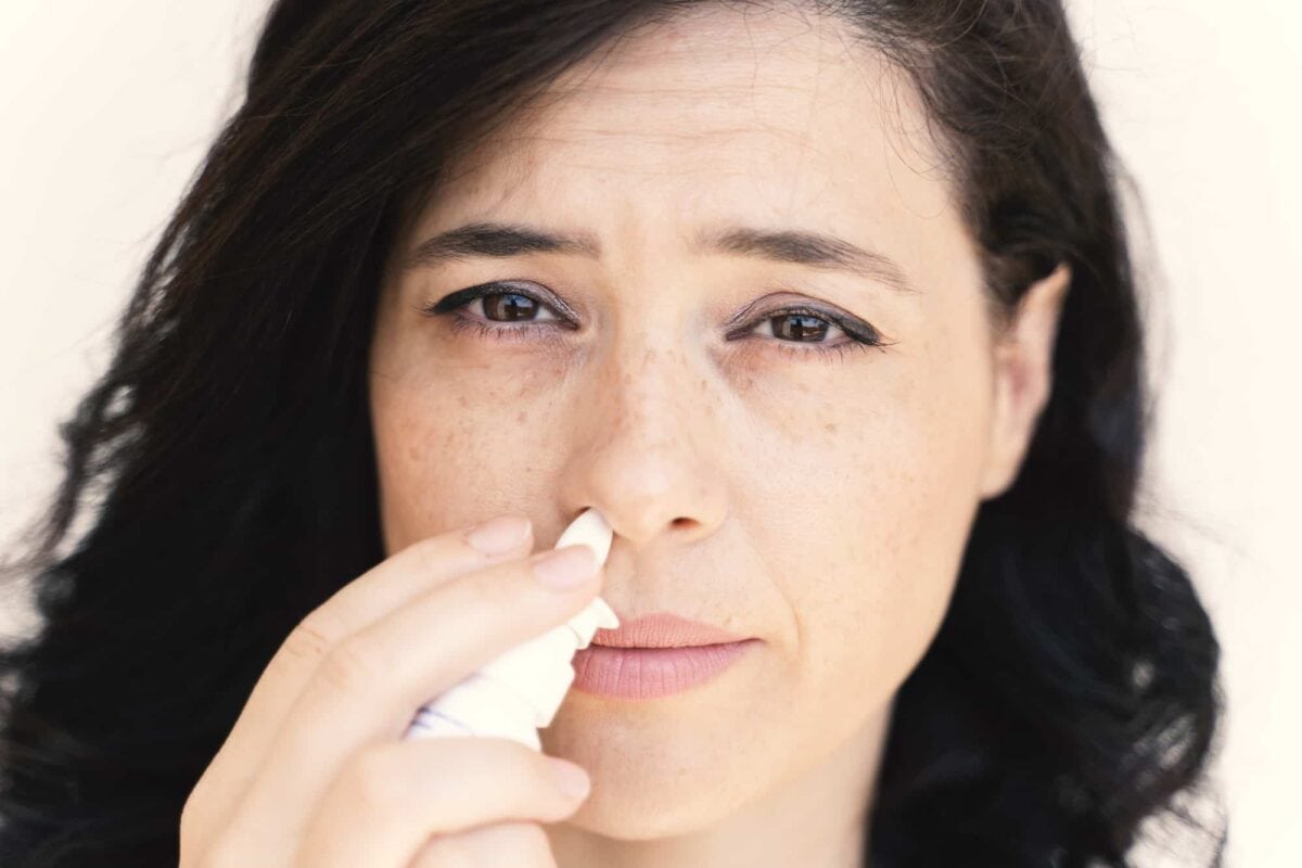 Woman with dark hair uses a nasal spray, holding the bottle to one nostril and looking directly at the camera.