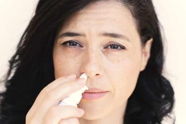 Woman with dark hair uses a nasal spray, holding the bottle to one nostril and looking directly at the camera.