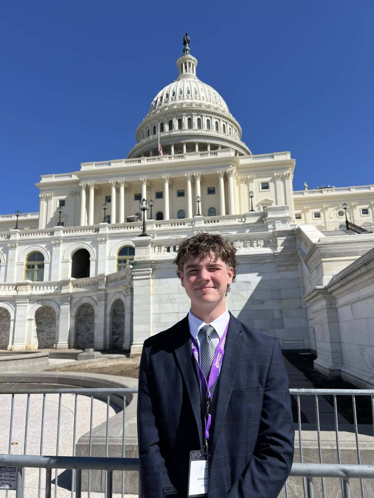 A young man in a suit stands outside the U.S. Capitol building on a sunny day, wearing an event badge and lanyard.
