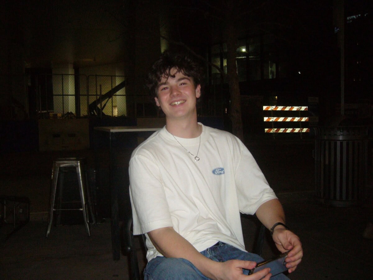 A young man with curly hair, wearing a white Ford t-shirt and jeans, sits and smiles at night in an urban outdoor setting.