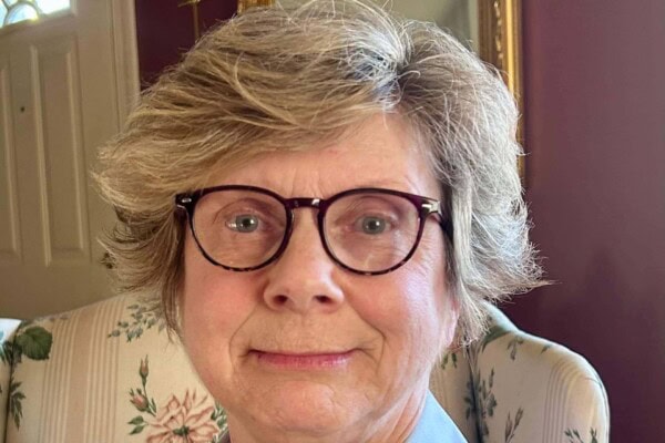 An older woman with short, light brown hair and glasses sits on a floral-patterned chair, looking at the camera.