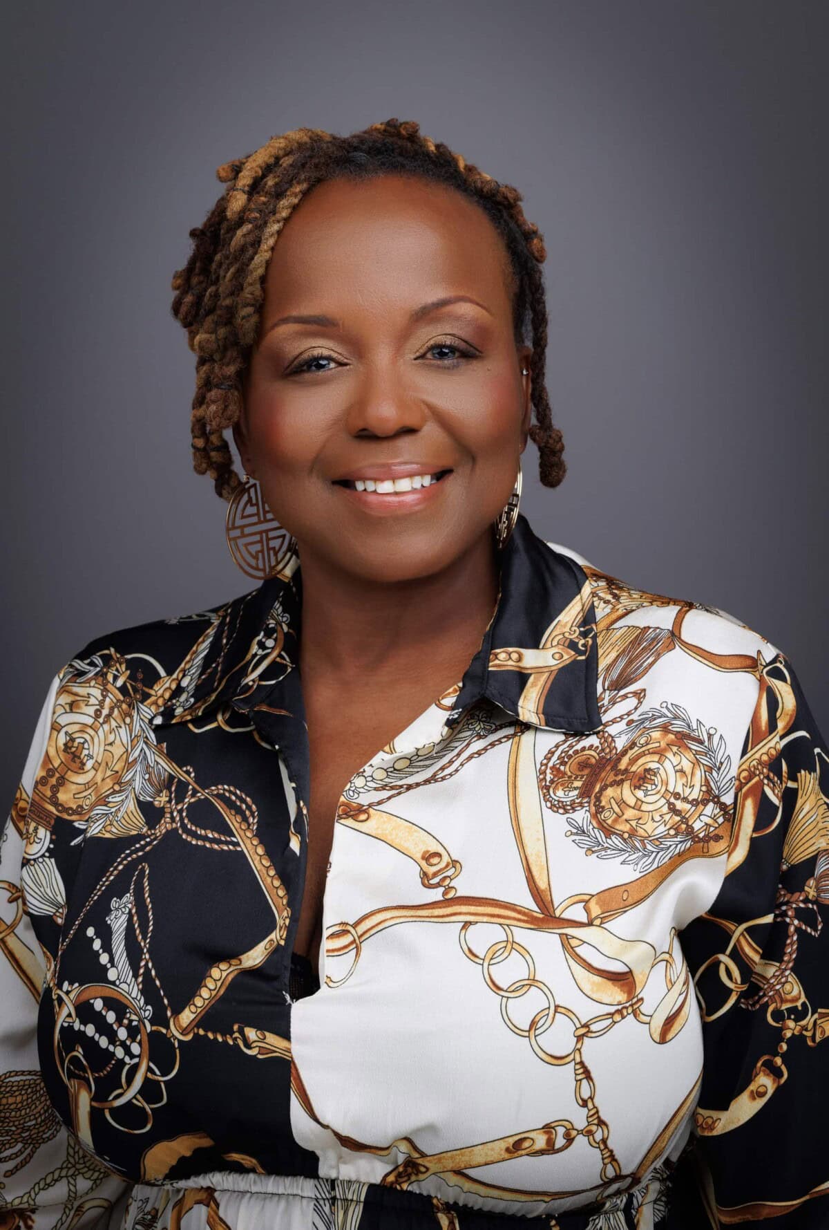 A woman with short, styled hair smiles at the camera, wearing a patterned shirt with gold chains and floral designs on a gray background.