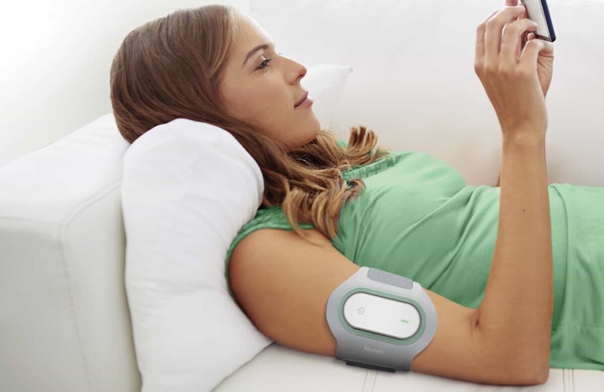 A woman lying on a couch uses her phone while wearing a gray electronic device strapped to her upper arm.