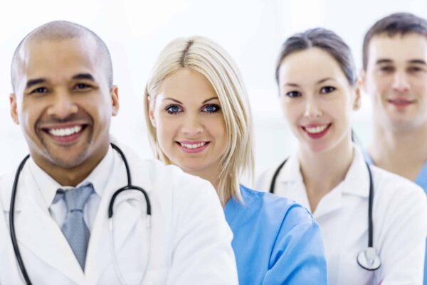 Four medical professionals, including men and women, stand in a row wearing lab coats and scrubs, smiling at the camera in a bright clinical setting.