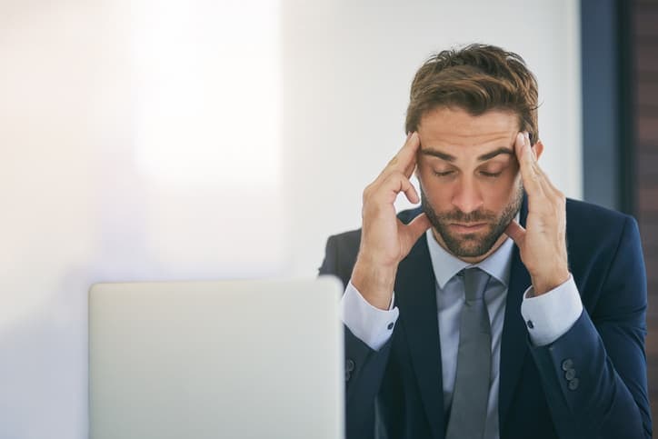 A man in a suit sits at a desk with a laptop, resting his head in his hands and appearing stressed or tired.